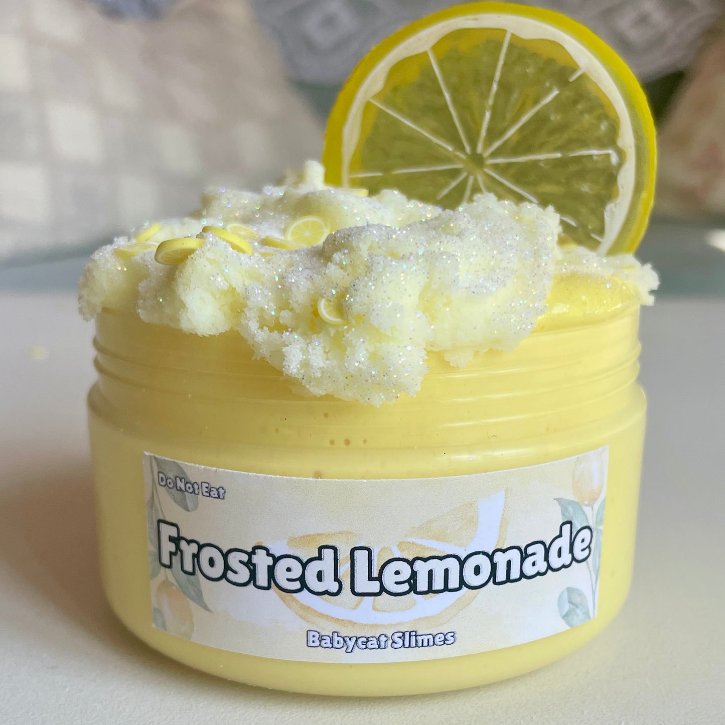 A jar is filled with yellow butter slime with a sparkly light yellow cloud slime on top. It's further topped with clay fimo lemons and a plastic lemon charm. The label reads "Frosted Lemonade, Babycat Slimes"