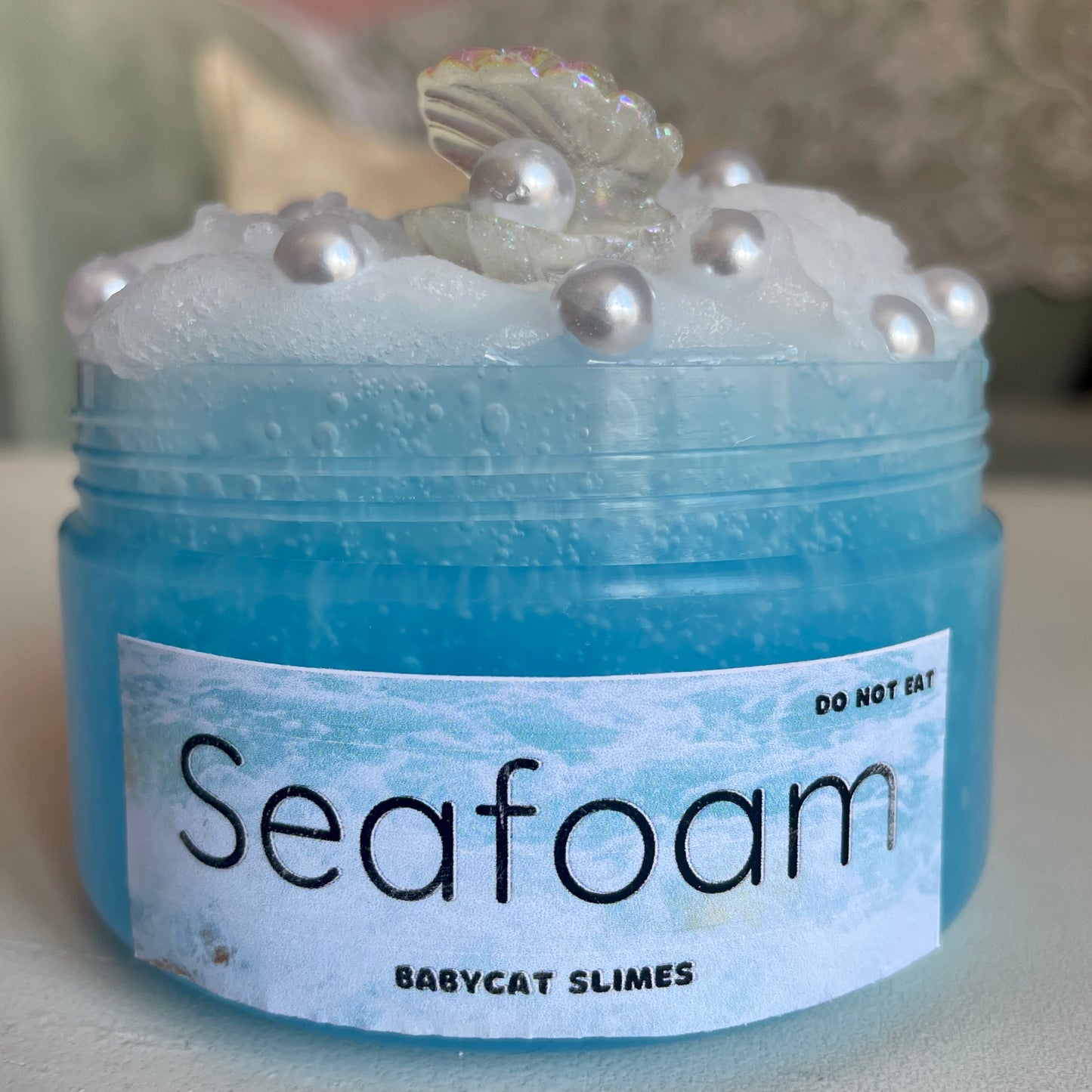 A transparent jar is filled with a light blue clear slime with a white icee slime on top. It's topped with pearl charms and an oyster + pearl charm. The label says "Seafoam - Babycat Slimes"