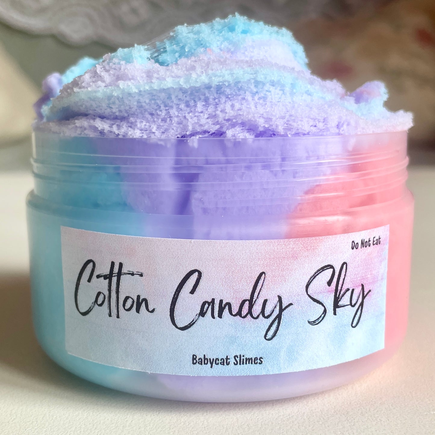 A container is filled with cloud slime in 3 different colors - light blue, purple, and pink, making a pastel sunset color combination which is swirled on the top. The label says "Cotton Candy Sky - Babycat Slimes"