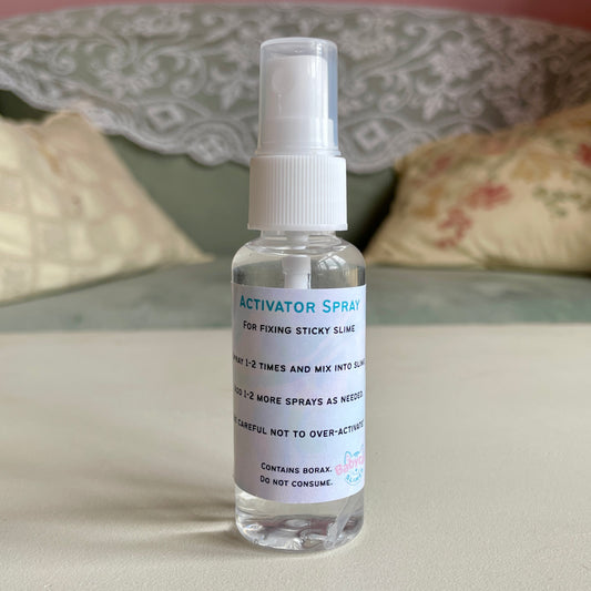 A 50ml clear spray bottle holds a clear liquid, which is a borax-based activator spray for fixing sticky slime. The label on it gives instructions for using the activator spray and the logo for Babycat Slimes.