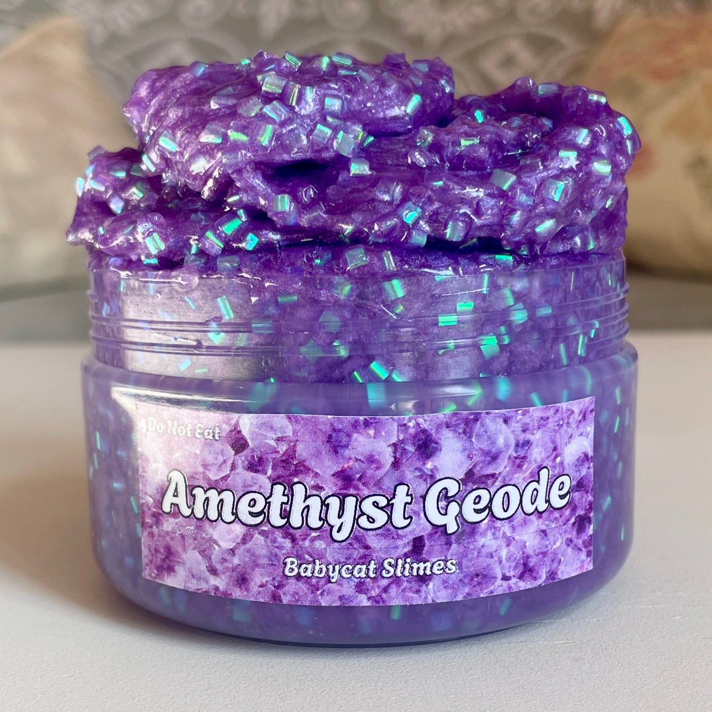 A transparent jar is overflowing with shimmering deep purple slime with bingsu beads. The label reads "Amethyst Geode by Babycat Slimes"