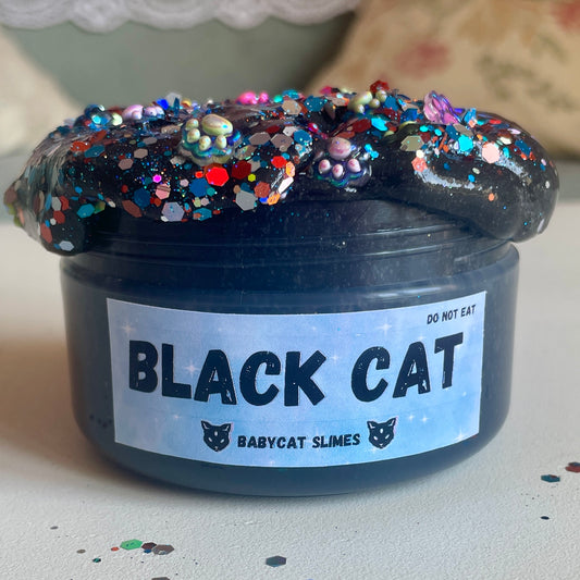 A jar is filled with black jelly slime and topped with rainbow glitter pieces and cat print charms. The label reads Black Cat Babycat Slimes.