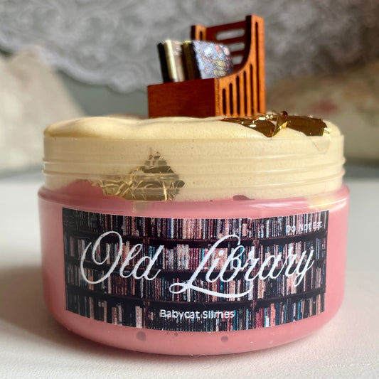 A container is filled with pink butter bingsu and beige WG jelly slime with pieces of gold leaf throughout. On top sits a bookshelf and tiny book charms. The label says "Old Library" and underneath, "Babycat Slimes".