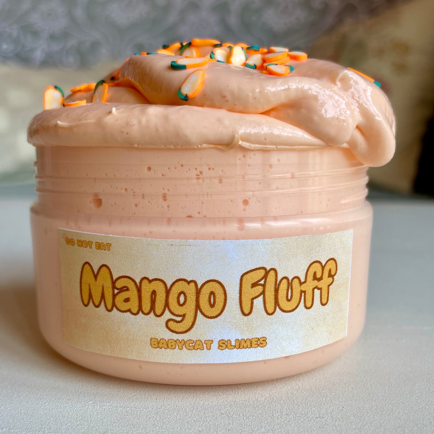 A jar is filled with orange butter slime with mango-shaped clay fimo chips on top. Label reads 'Mango Fluff' 'Babycat Slimes'