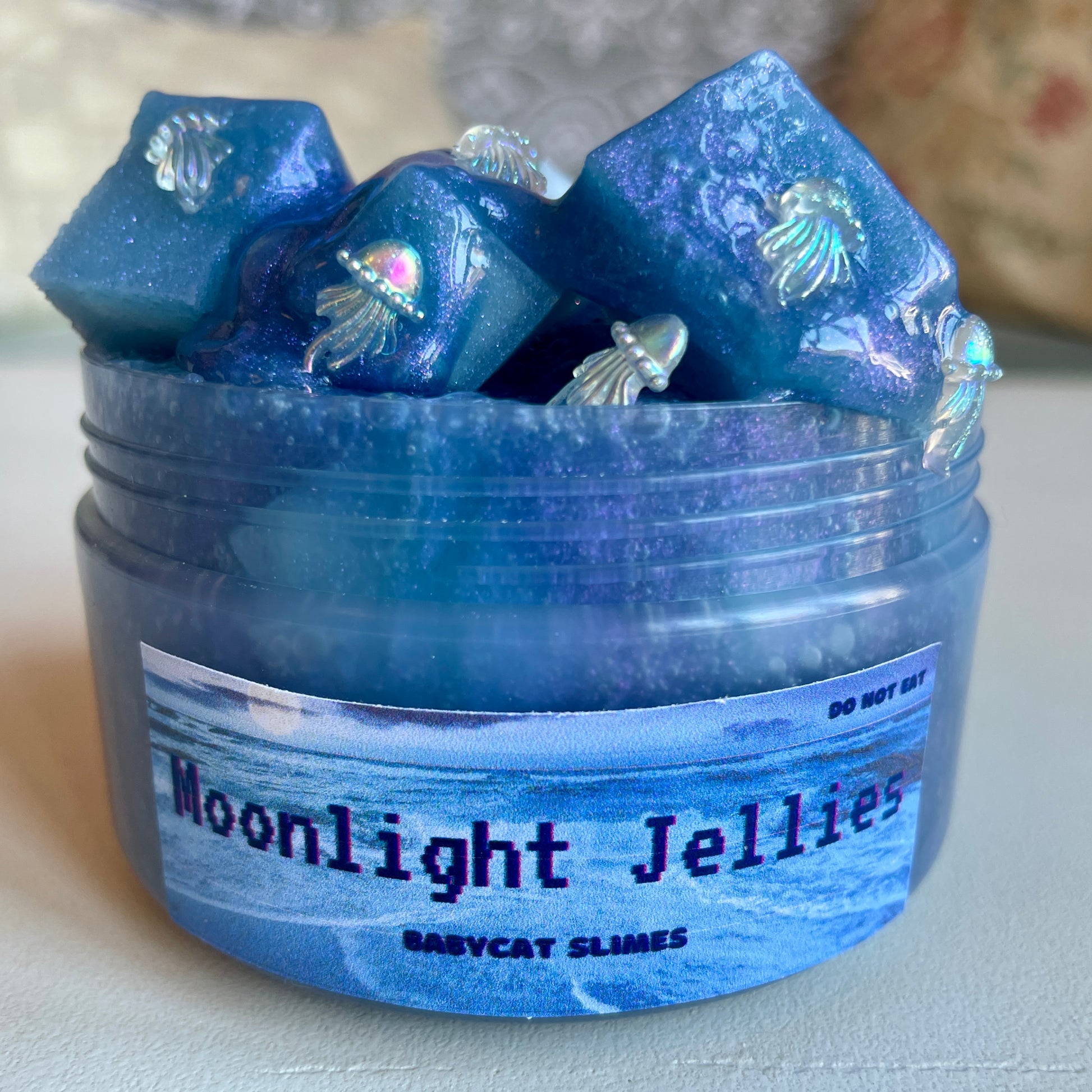 A jar is filled with a shimmering blue colorshift slime with jelly success and iridescent jellyfish charms. The label says Moonlight Jellies Babycat Slimes