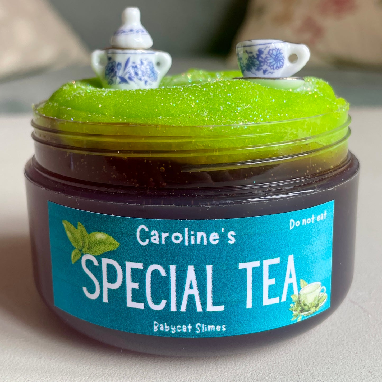 A transparent jar is filled with a tea-coloured clear slime and topped with a sparkly green icee slime. There are miniature tea set charms on top. The label on the slime reads "Caroline's Special Tea - Babycat Slimes"