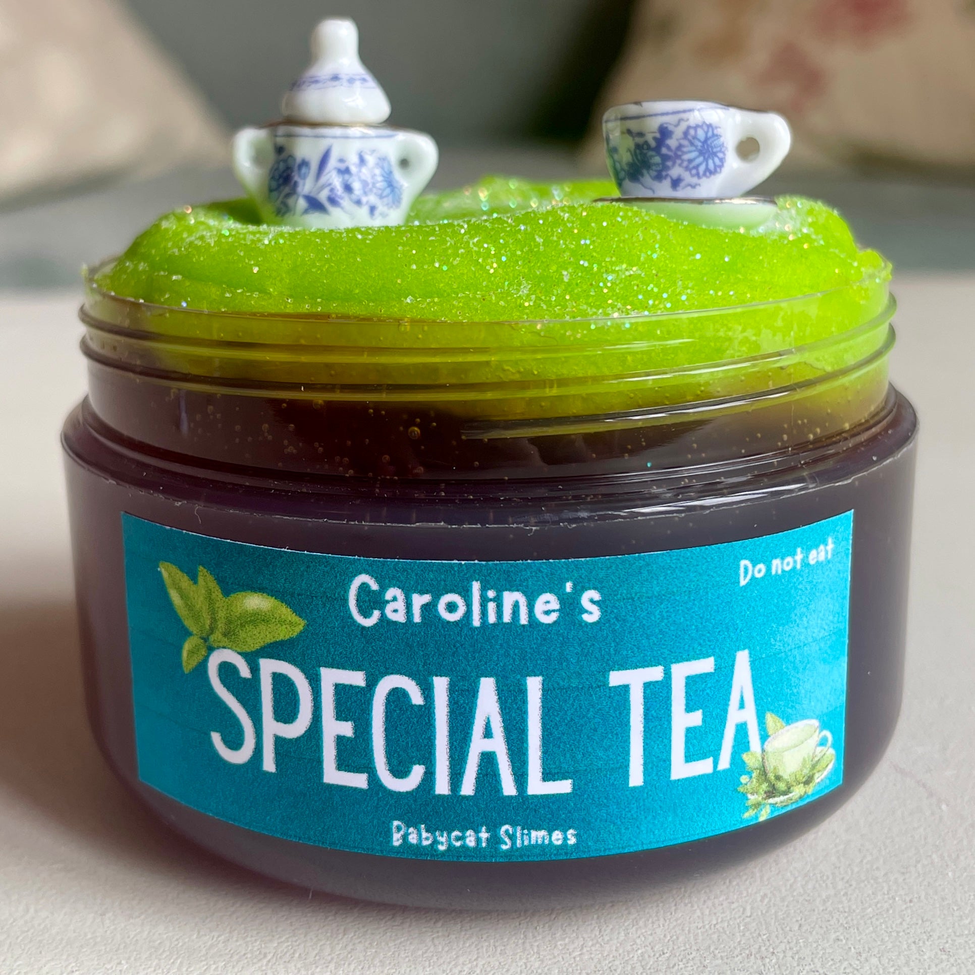 A transparent jar is filled with a tea-coloured clear slime and topped with a sparkly green icee slime. There are miniature tea set charms on top. The label on the slime reads "Caroline's Special Tea - Babycat Slimes"