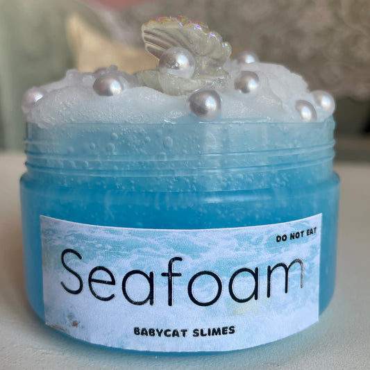 A transparent jar is filled with a light blue clear slime with a white icee slime on top. It's topped with pearl charms and an oyster + pearl charm. The label says "Seafoam - Babycat Slimes"
