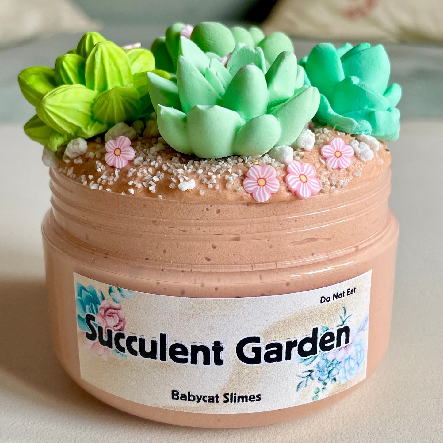 A container is filled with a tan cloud creme slime, and topped with sand, perlite rocks, pink flower clay fimo pieces, and 4 squishable clay succulent plants. The label says "Succulent Garden - Babycat Slimes"