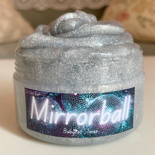 A jar is filled with glittery silver coated clear slime with rainbow sparkles. The label says Mirrorball Babycat Slimes