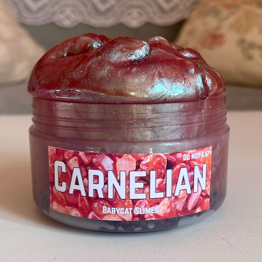 A jar is filled with shimmering colorshift red coated clear slime with black rocks at the bottom. The label reads "Carnelian by Babycat Slimes"