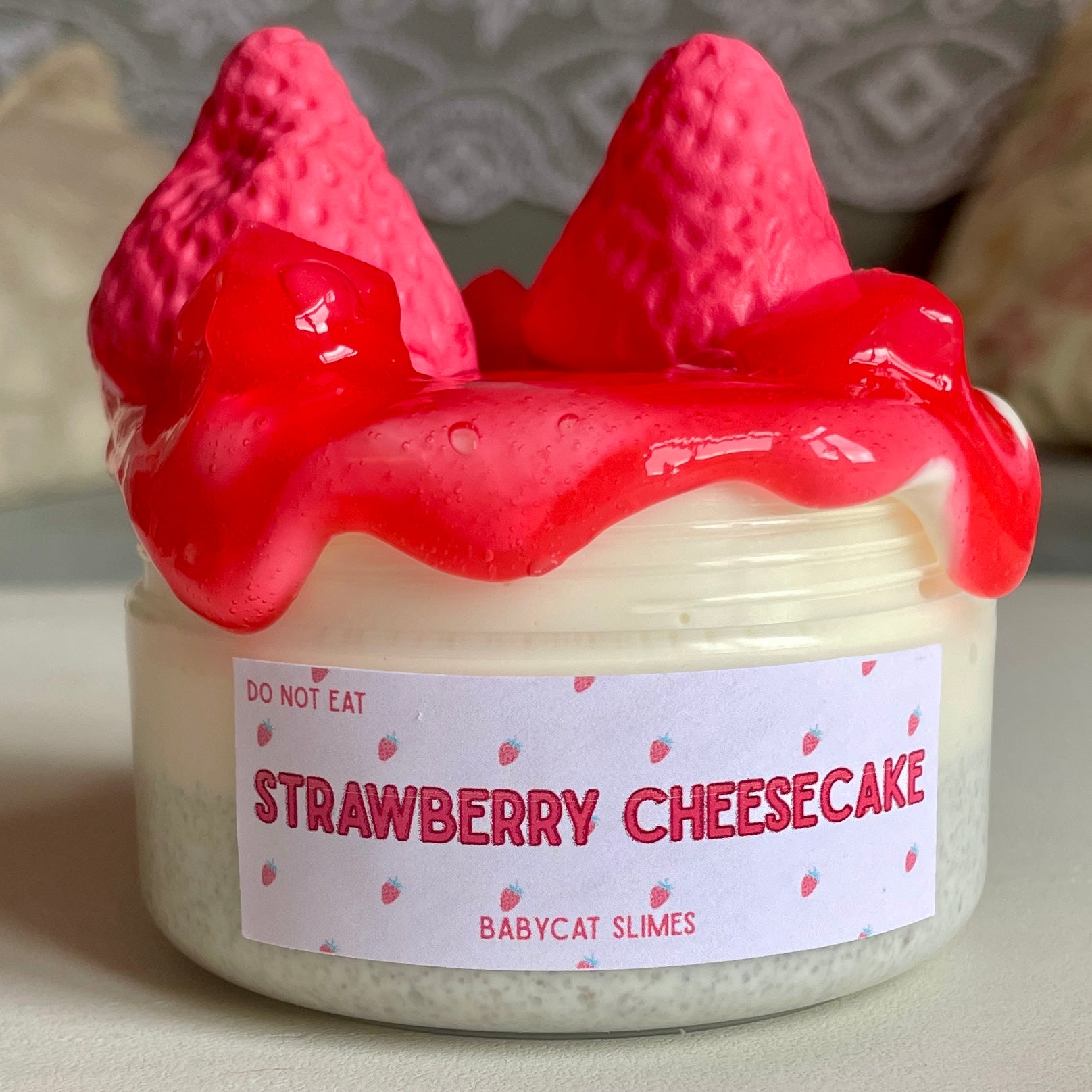 A jar is filled with a light yellow slime with sand crust at the bottom. It's topped with a red slime with jelly cubes, and on top of that are two red squishable clay strawberries. The label reads "Strawberry Cheesecake by Babycat Slimes".