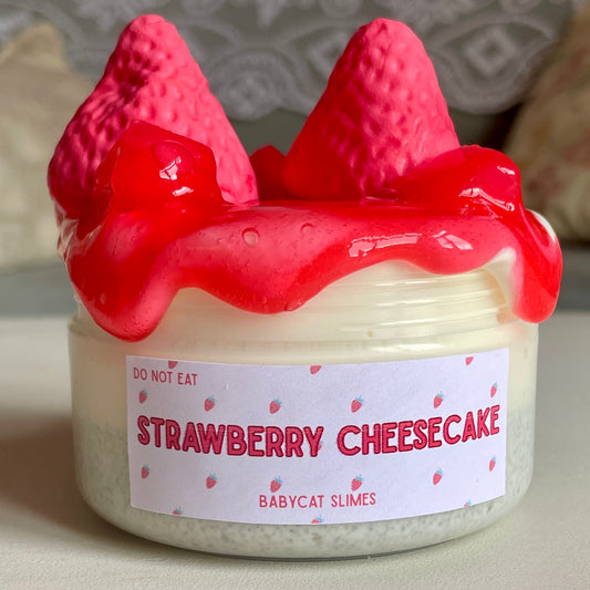 A jar is filled with a light yellow slime with sand crust at the bottom. It's topped with a red slime with jelly cubes, and on top of that are two red squishable clay strawberries. The label reads "Strawberry Cheesecake by Babycat Slimes".