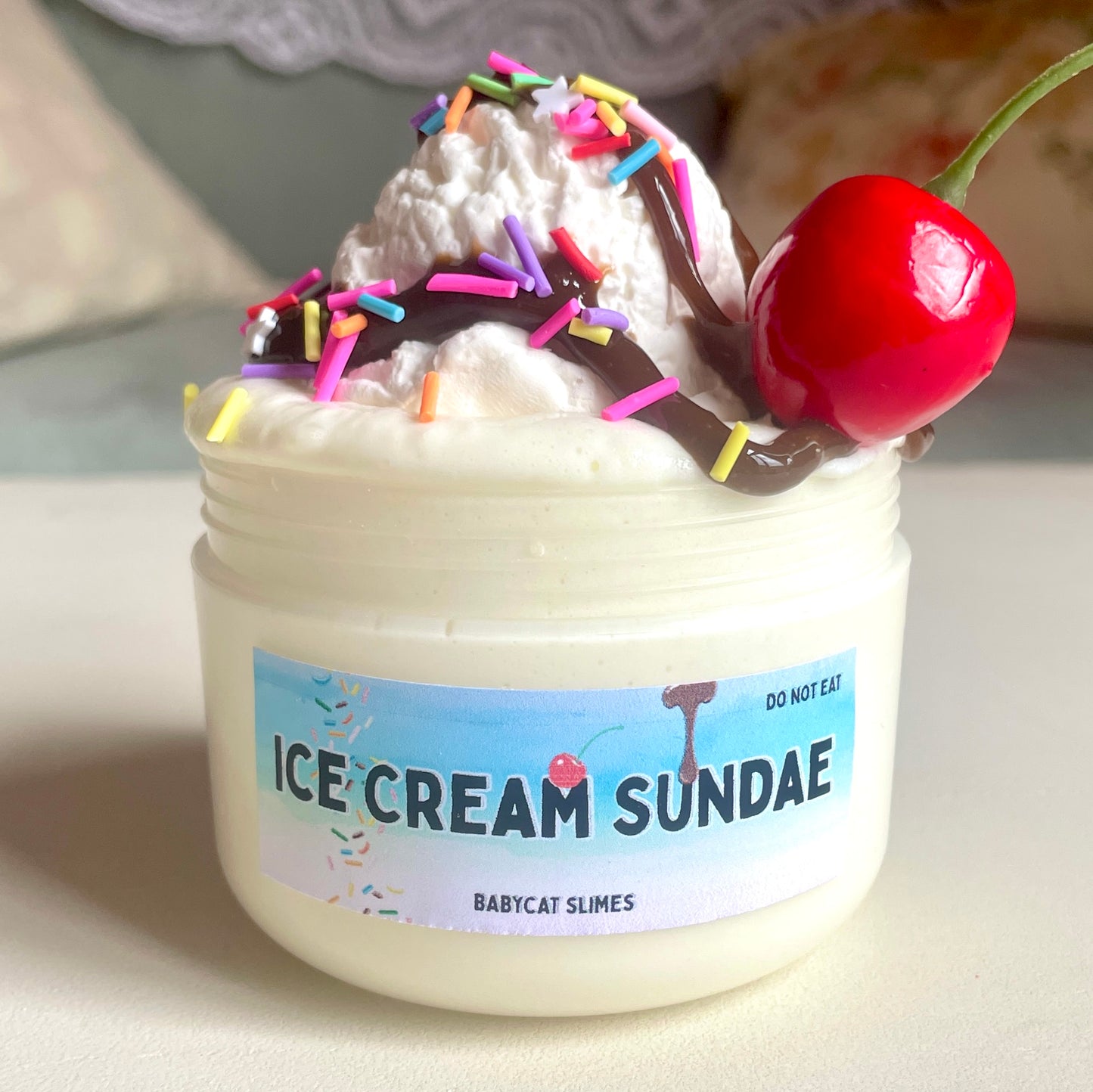 A jar is filled with white signature "soft serve" texture slime with a squishable clay ice cream scoop on top, and a brown slime resembling chocolate sauce drizzled over it. It's topped with clay fimo sprinkles and a cherry charm. The label reads "Ice Cream Sundae - Babycat Slimes"