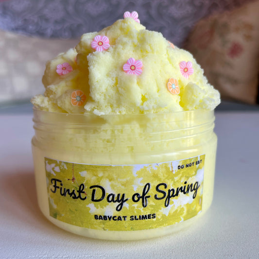 A jar is filled with a fluffy yellow cloud slime with orange and pink flower fimo pieces. The label says "First Day of Spring by Babycat Slimes"