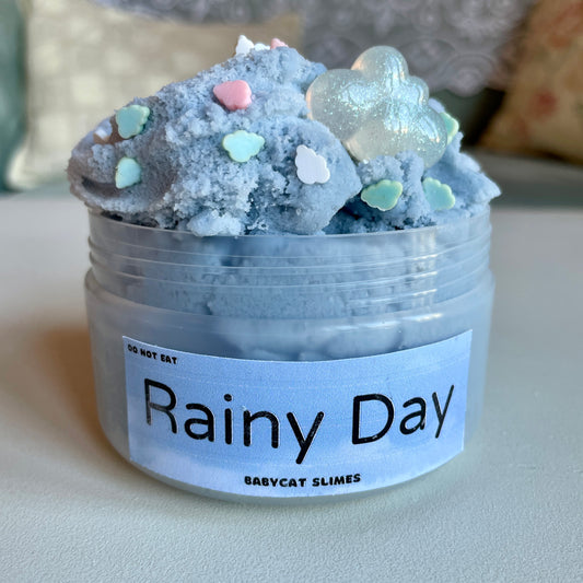 A container is filled with blue-grey fluffy cloud slime with blue, pink, and white fimo clay clouds and a sparkling resin cloud charm. The label says Rainy Day by Babycat Slimes.
