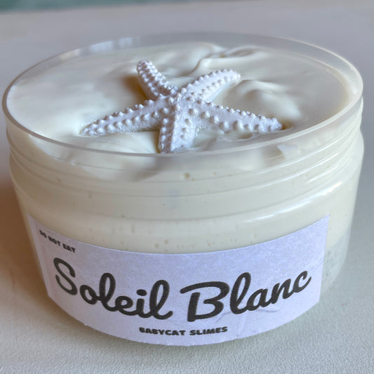 A jar is filled with creamy white glossy slime with a layer of sand at the bottom. There is a starfish charm on top. The label says Soleil Blanc by Babycat Slimes