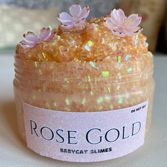 A container is filled with shimmering gold bingsu slime with 3 pink and gold flower charms. Label reads Rose Gold Babycat Slimes.