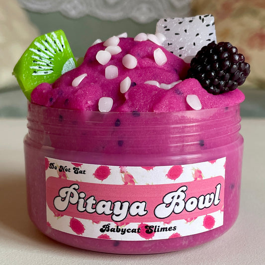 A jar is filled with a purpleish pink icee slime with little clay pieces resembling seeds throughout. On top are small wax pieces meant to look like coconut flakes. There are 3 fruit charms on top. The label says "Pitaya Bowl, Babycat Slimes".