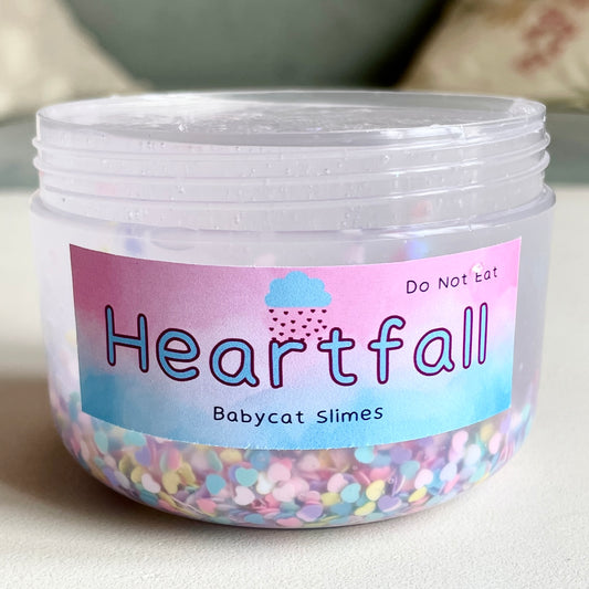 A transparent container is filled with clear slime, with tiny rainbow heart sequins on the bottom. The label says "Heartfall" in big letters and "Babycat Slimes" underneath.