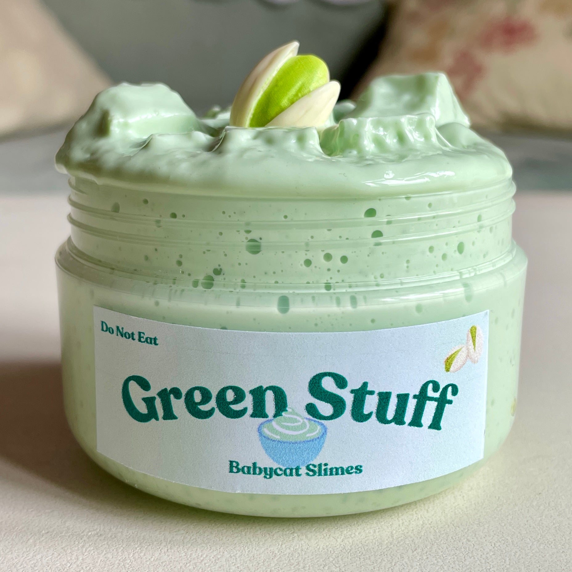 A container is filled with a fluffy light green slay slime with slushee beads and jelly cubes, and a pistachio charm on top. The label says "Green Stuff" and underneath, "Babycat Slimes".