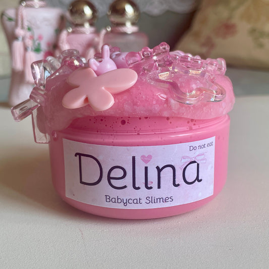 A jar is filled with bright pink slay slime and topped with a sparkling clear slime. On top are several coquette and kawaii beads, shaped like bows, butterflies, stars, bunnies, hearts, and other cute kawaii things. The label says Delina Babycat Slimes