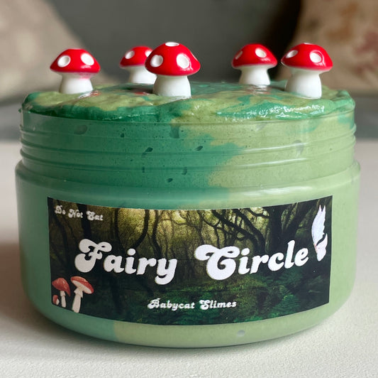 A transparent jar is filled with two-toned green butter slime with perlite. On top are 5 red mushroom charms. The label reads "Fairy Circle - Babycat Slimes"