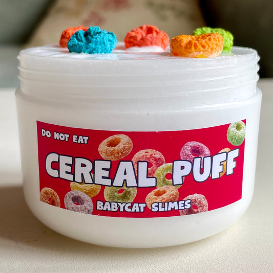 A transparent container is filled with white "WG jelly" slime with rainbow clay cereal pieces on top. The label says "Cereal Puff" in big letters, with "Babycat Slimes" underneath.